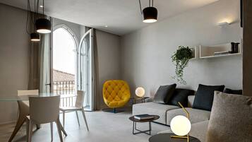 Una sala moderna con una mesa de comedor de vidrio, un sillón amarillo, un sofá y una mesa de centro. Hay lámparas colgantes, una planta y una estantería.