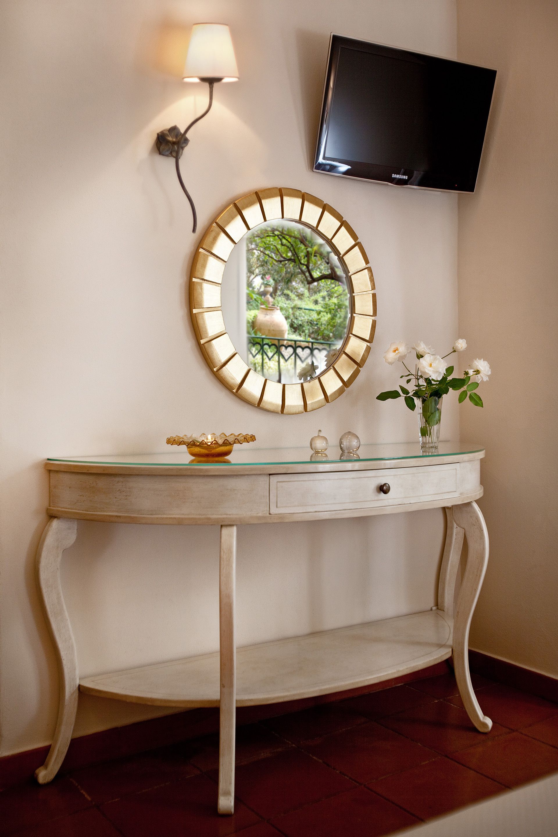 Een witte console tafel met een ronde spiegel, een klein vaasje met witte bloemen, een tv aan de muur en een wandlamp.