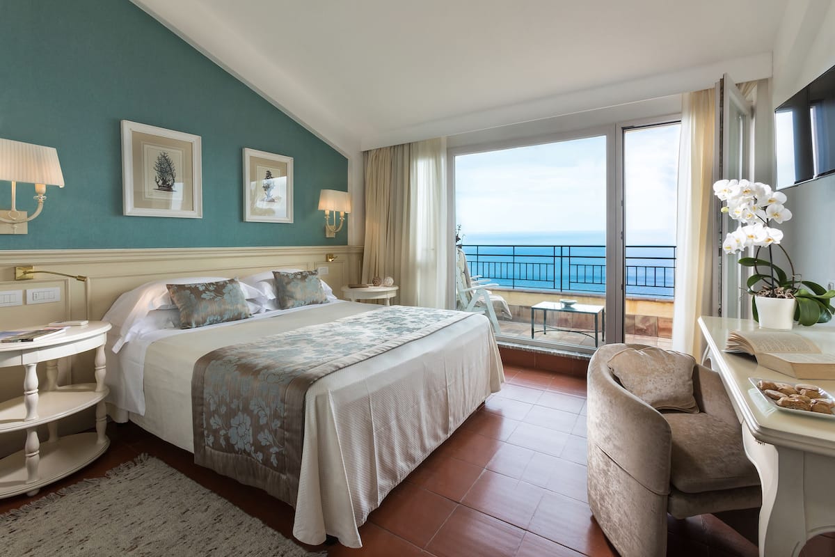 Een hotelkamer met een bed, een bureau, een stoel, een balkon met uitzicht op zee en een groot raam.