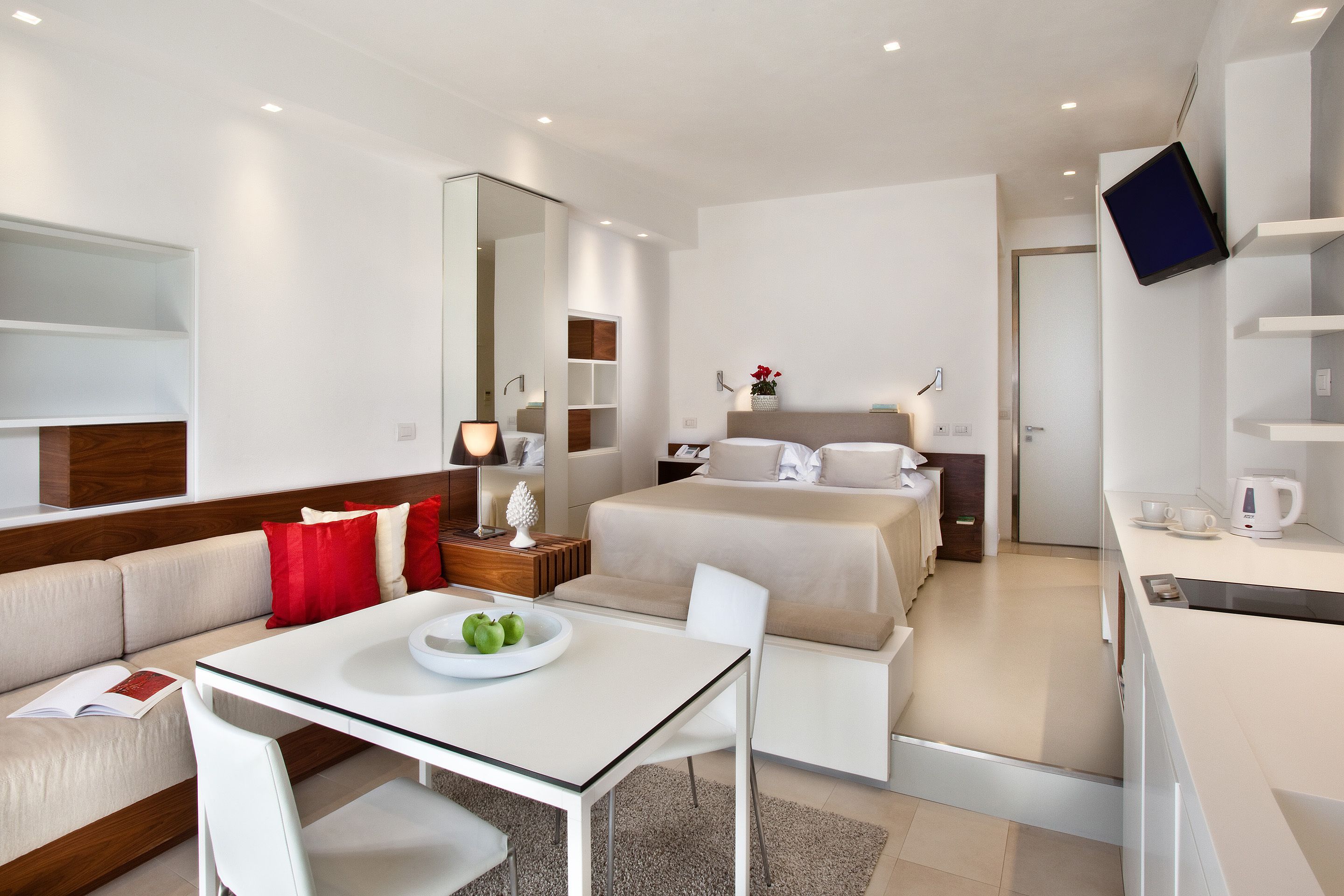 A modern hotel room with a sofa, a dining table, a bed, a TV mounted on the wall, and a kitchen area with a coffee maker.