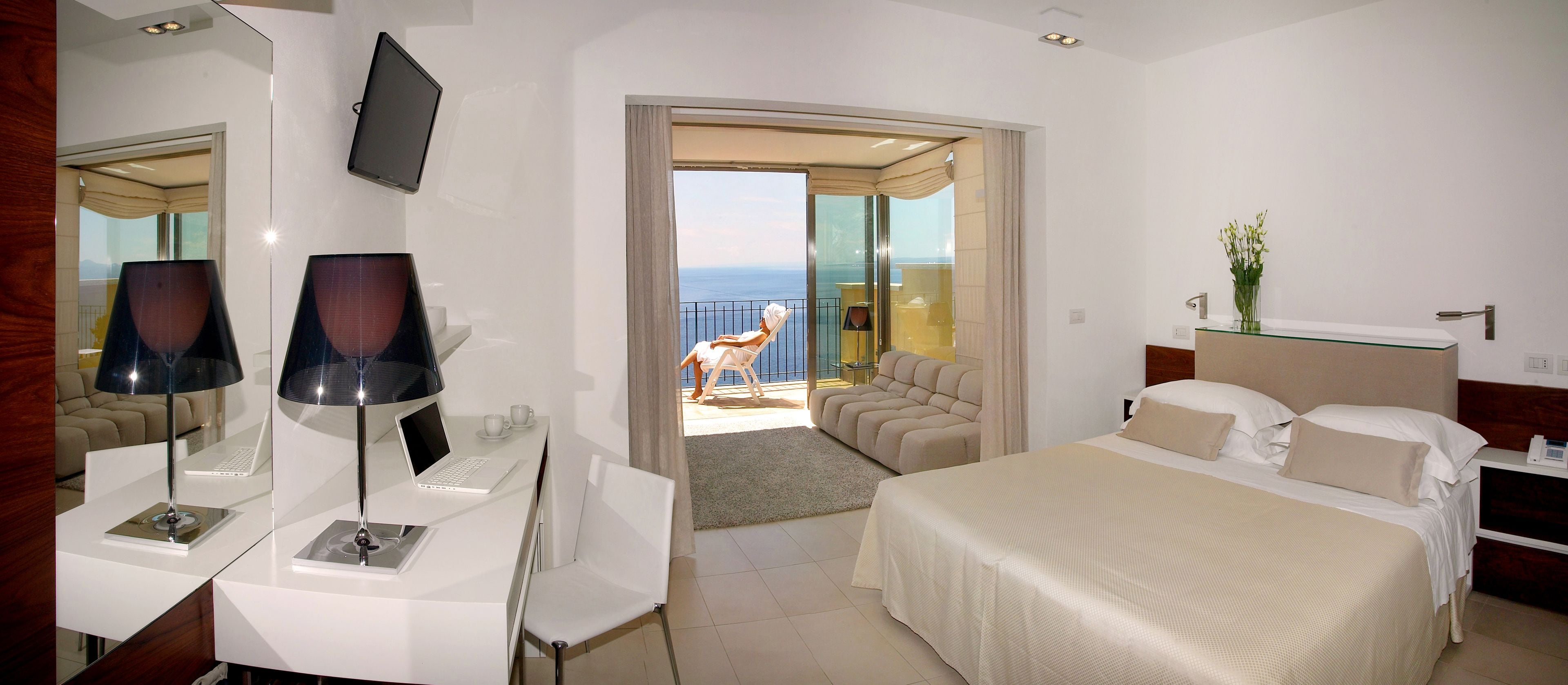 Camera d'albergo con un letto, una scrivania con un portatile, una sedia, una televisione e vista sul mare.