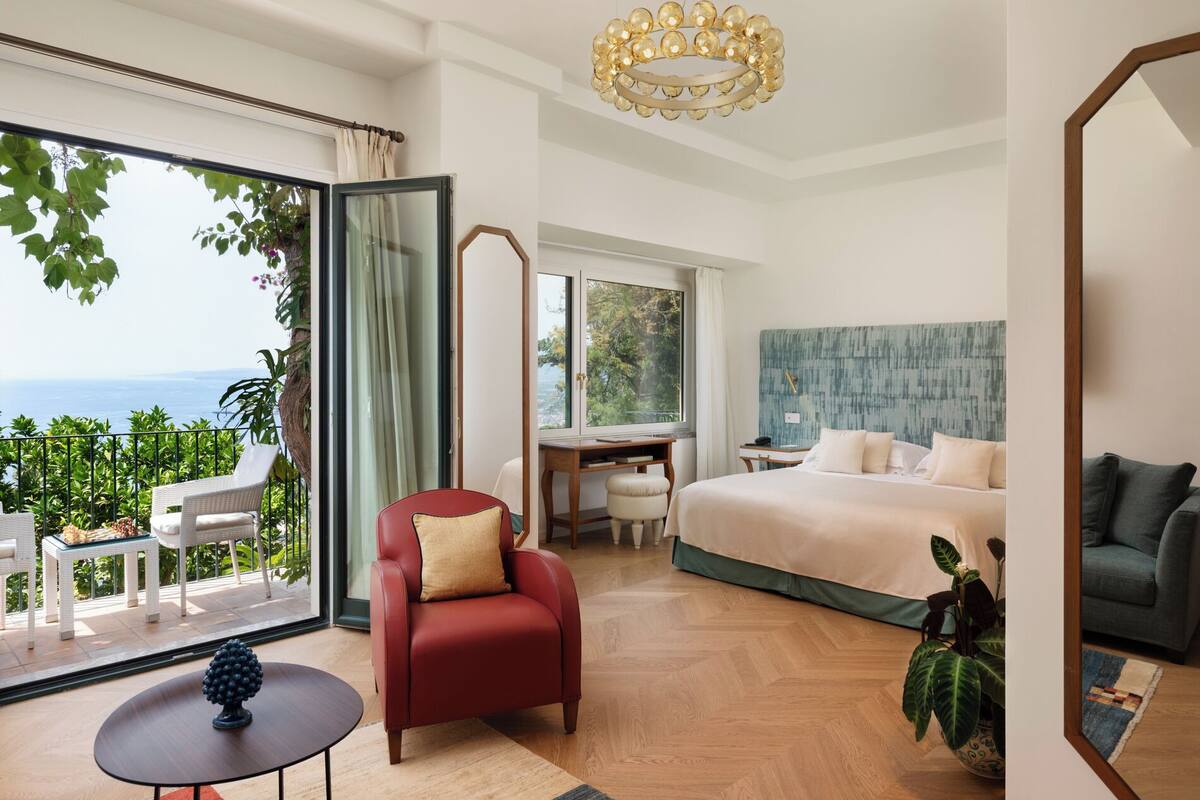 Een hotelkamer met een bed, een bureau, een rode fauteuil, een klein tafeltje en uitzicht op zee.