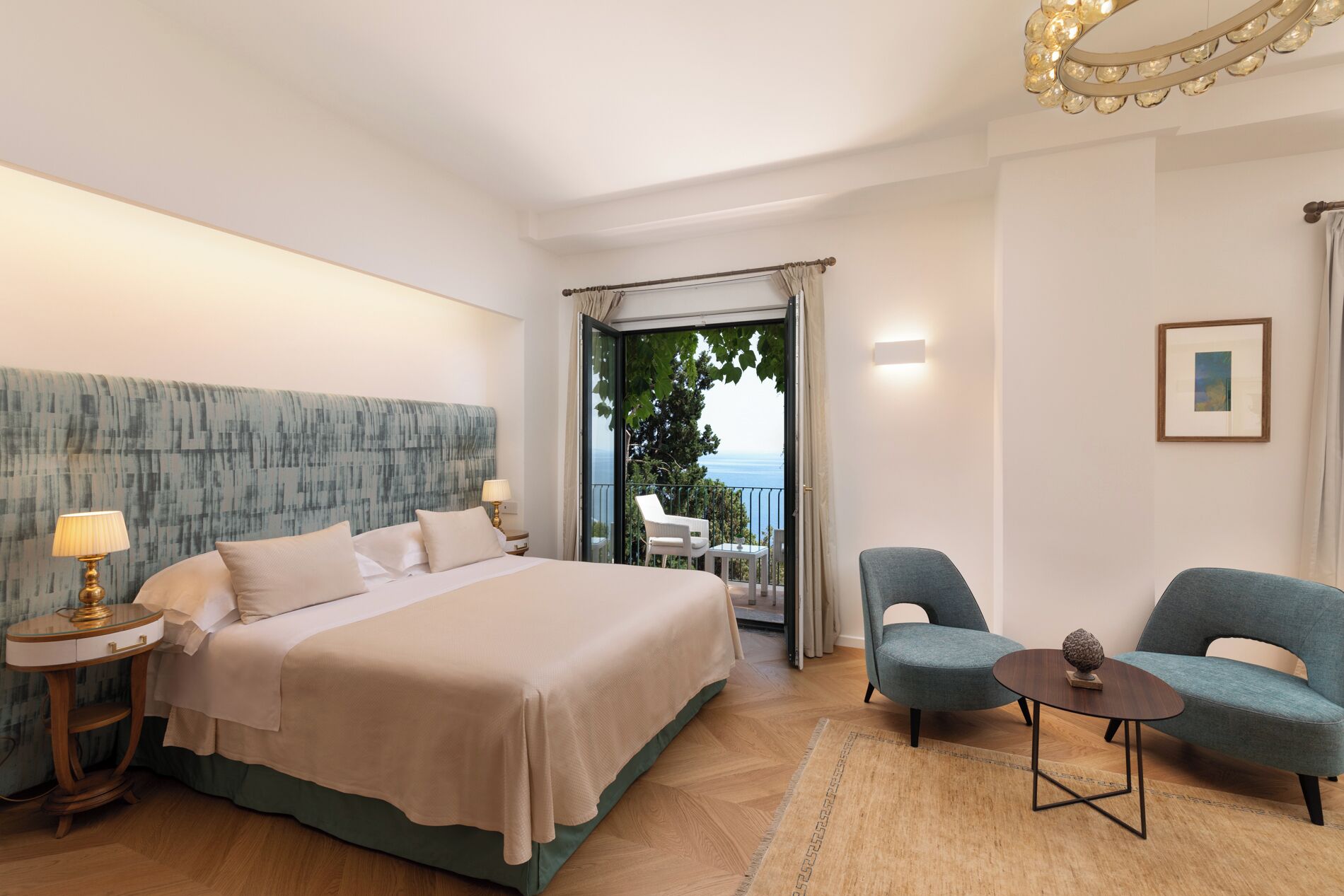 Camera d'albergo con un ampio letto, due poltrone, un tavolino, un balcone con vista e un lampadario.