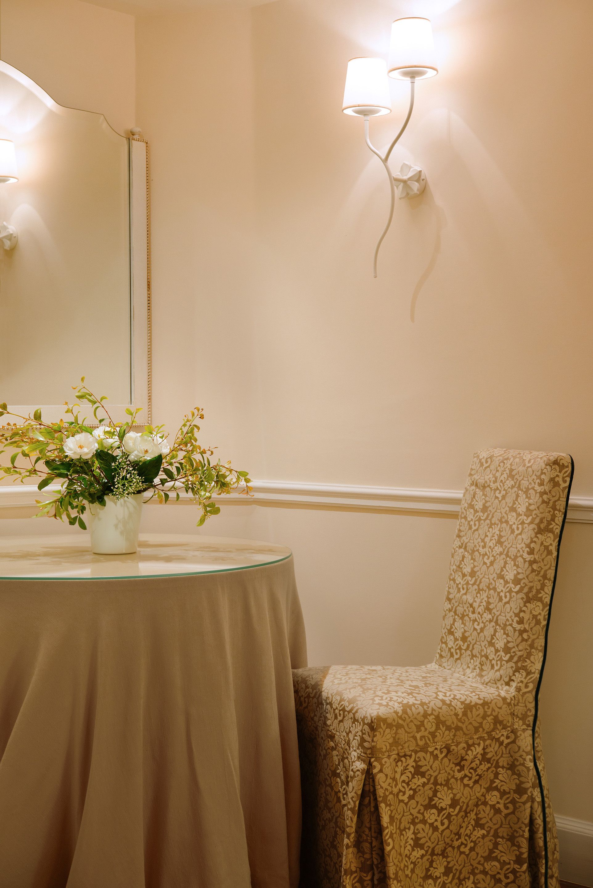 Ein Zimmer mit einem runden Tisch, einem Stuhl, einer Vase mit Blumen und Wandleuchten.