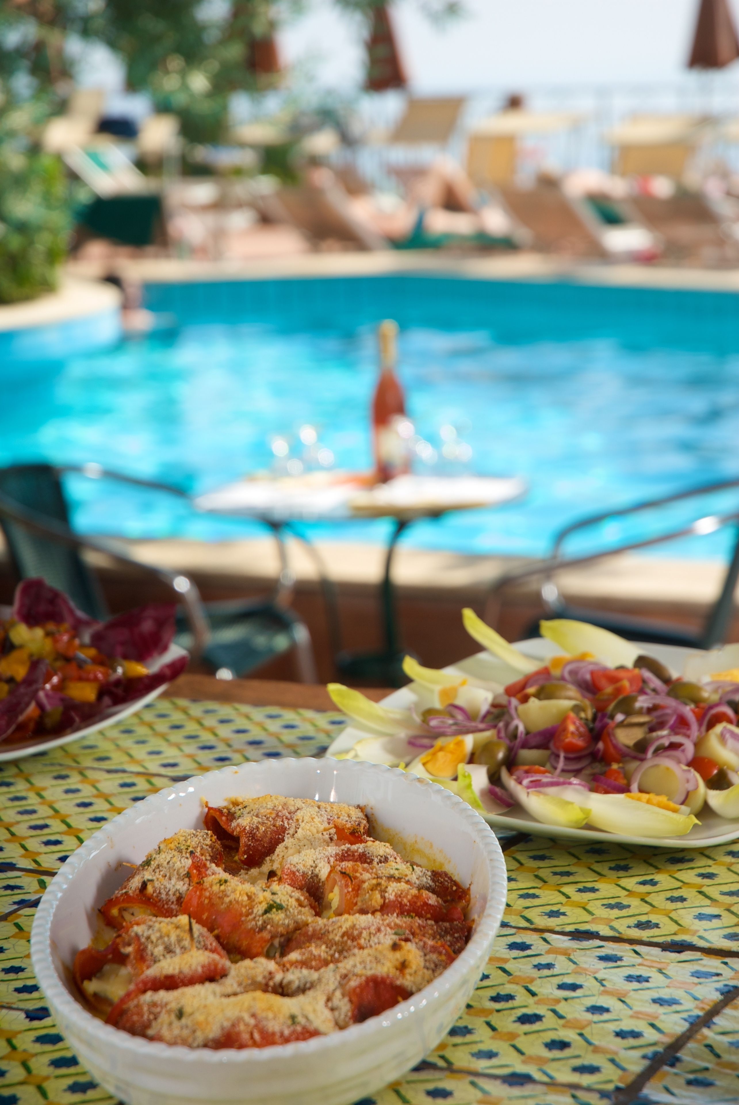 A poolside dining area with a table set for two, featuring a bowl of stuffed peppers and a plate of salad.