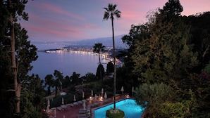 Un espace piscine doté de chaises longues et de parasols, entouré d’arbres et offrant une vue sur un plan d’eau, avec les lumières de la ville scintillantes au loin.