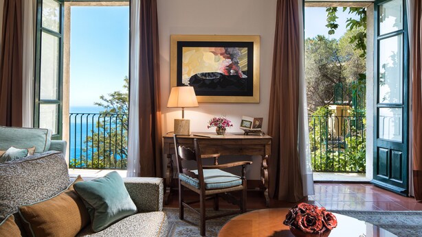 Une chambre avec vue sur la mer, un bureau avec une lampe, une chaise et une table ornée d’un vase de fleurs.