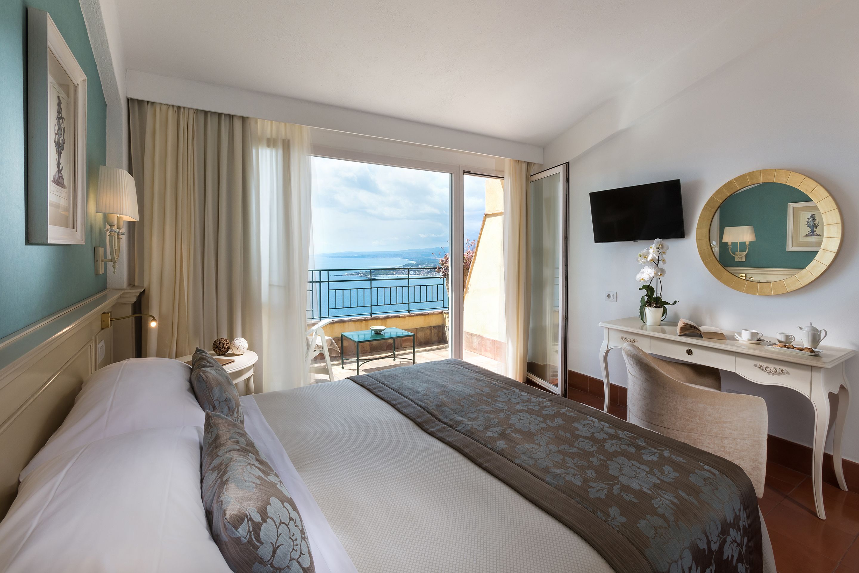 A hotel room with a large bed, a desk, a chair, a TV, and a balcony with a view of the sea.