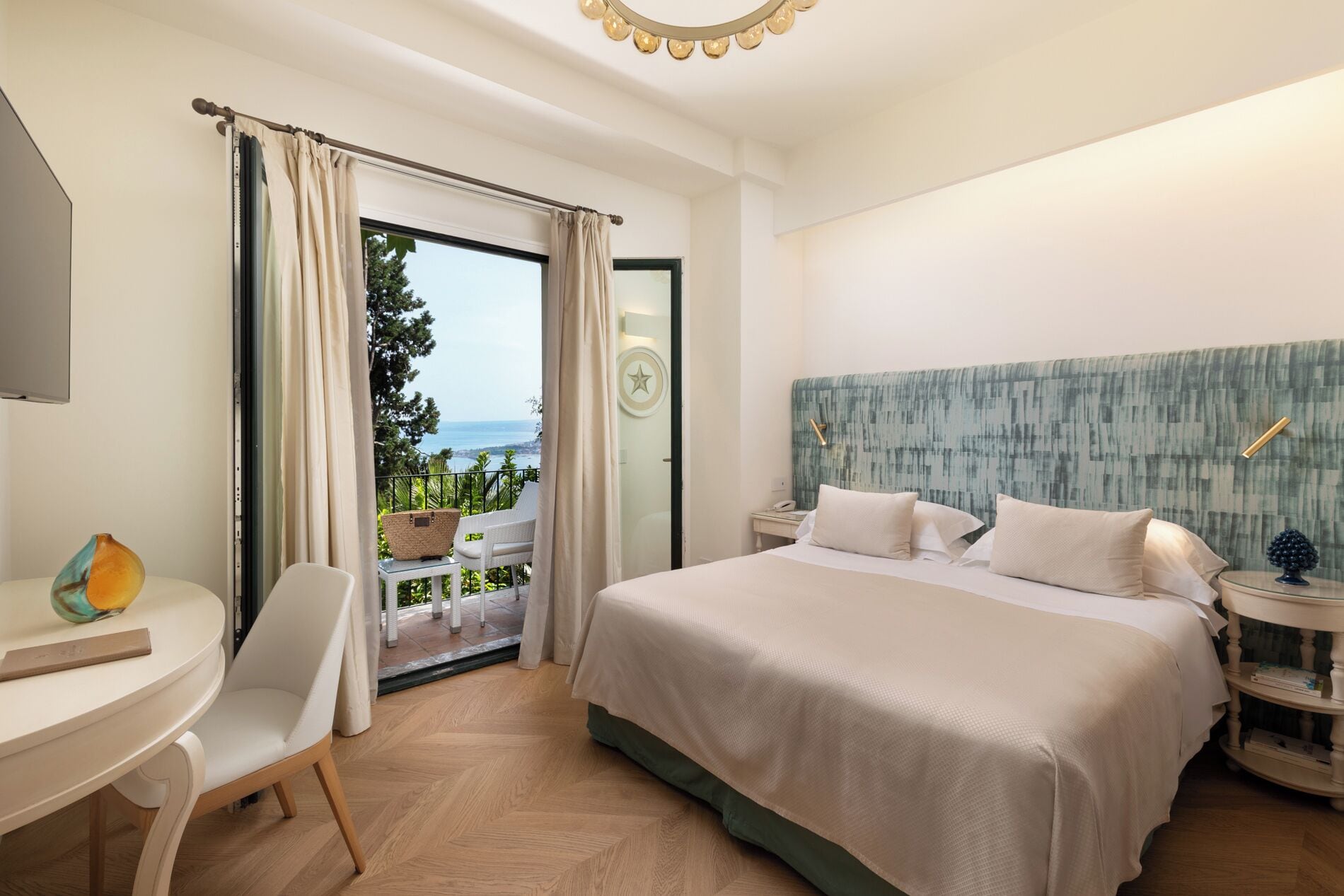 Une chambre d’hôtel avec un grand lit, un bureau, une chaise, une table de chevet et une vue sur la mer depuis un balcon.