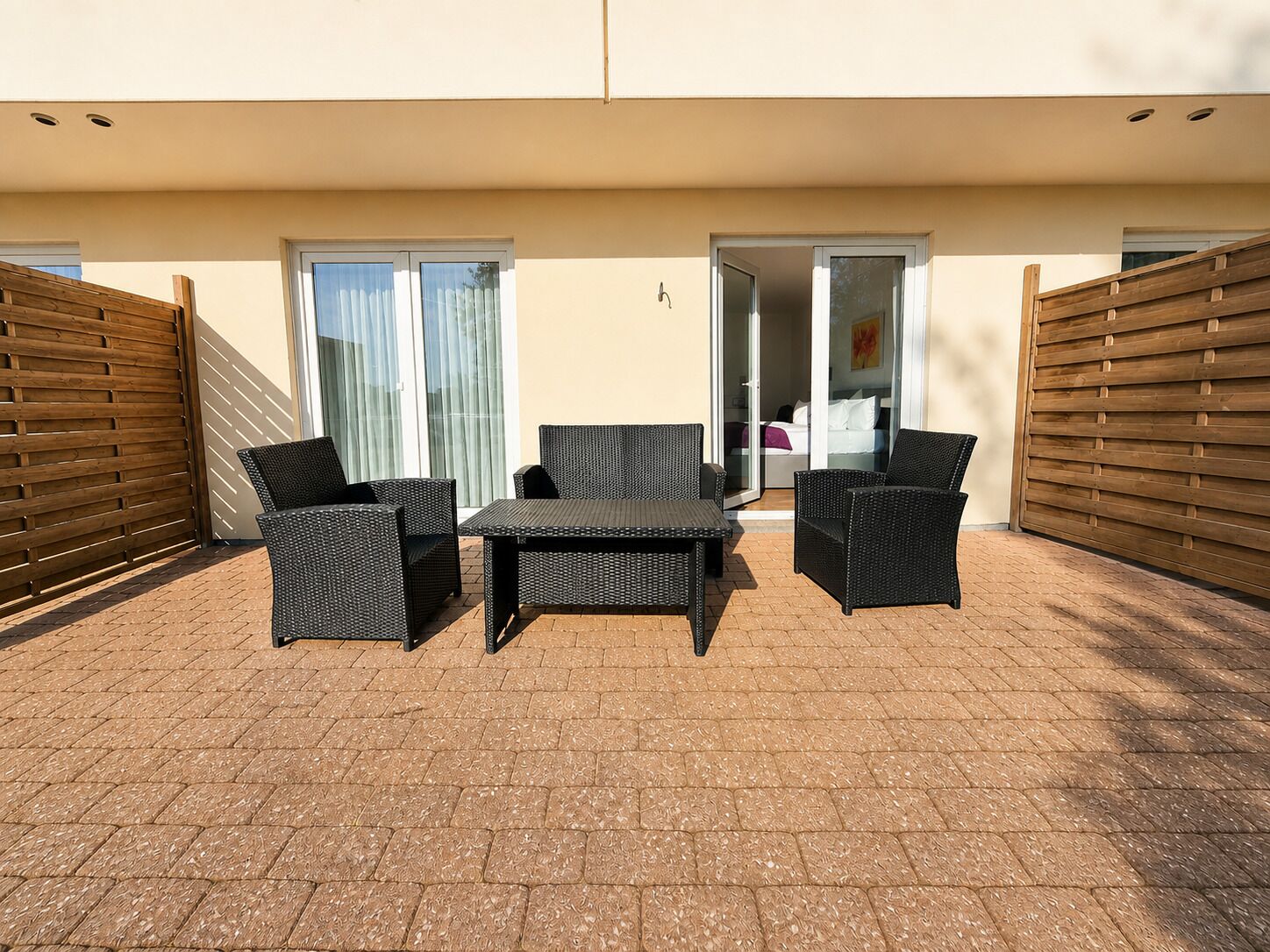 Un patio al aire libre con mobiliario de mimbre, una mesa y un ventanal corredera que da a un dormitorio.