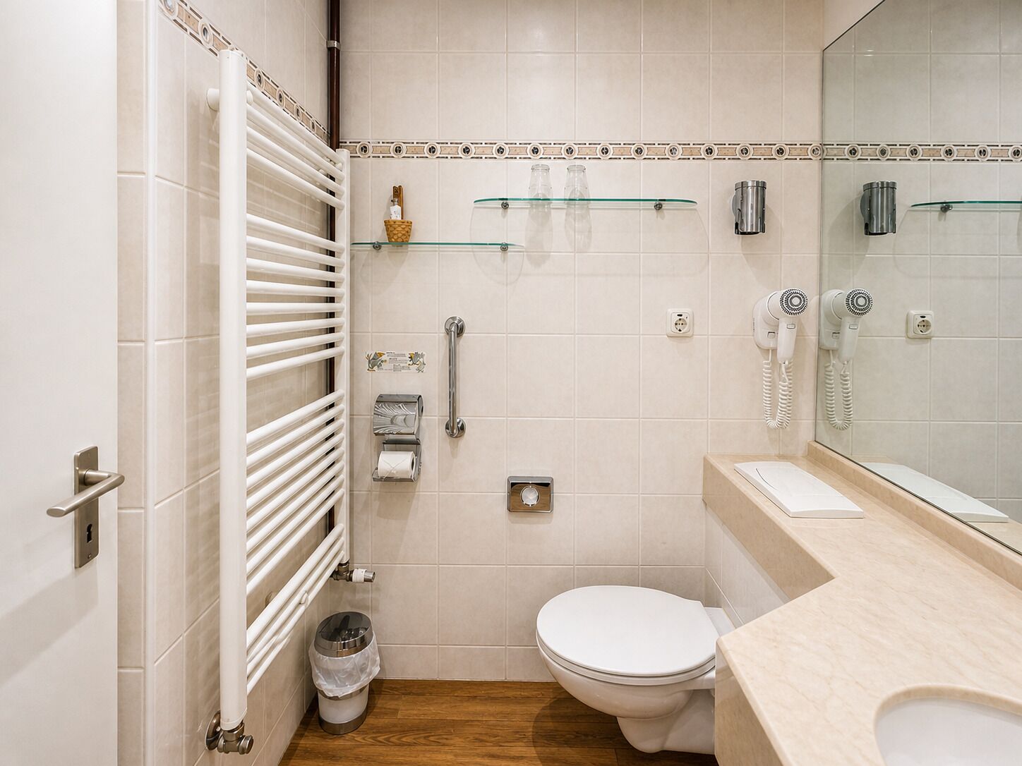 Un baño con inodoro, repisa de vidrio, secador de manos, cubo de basura y toallero.