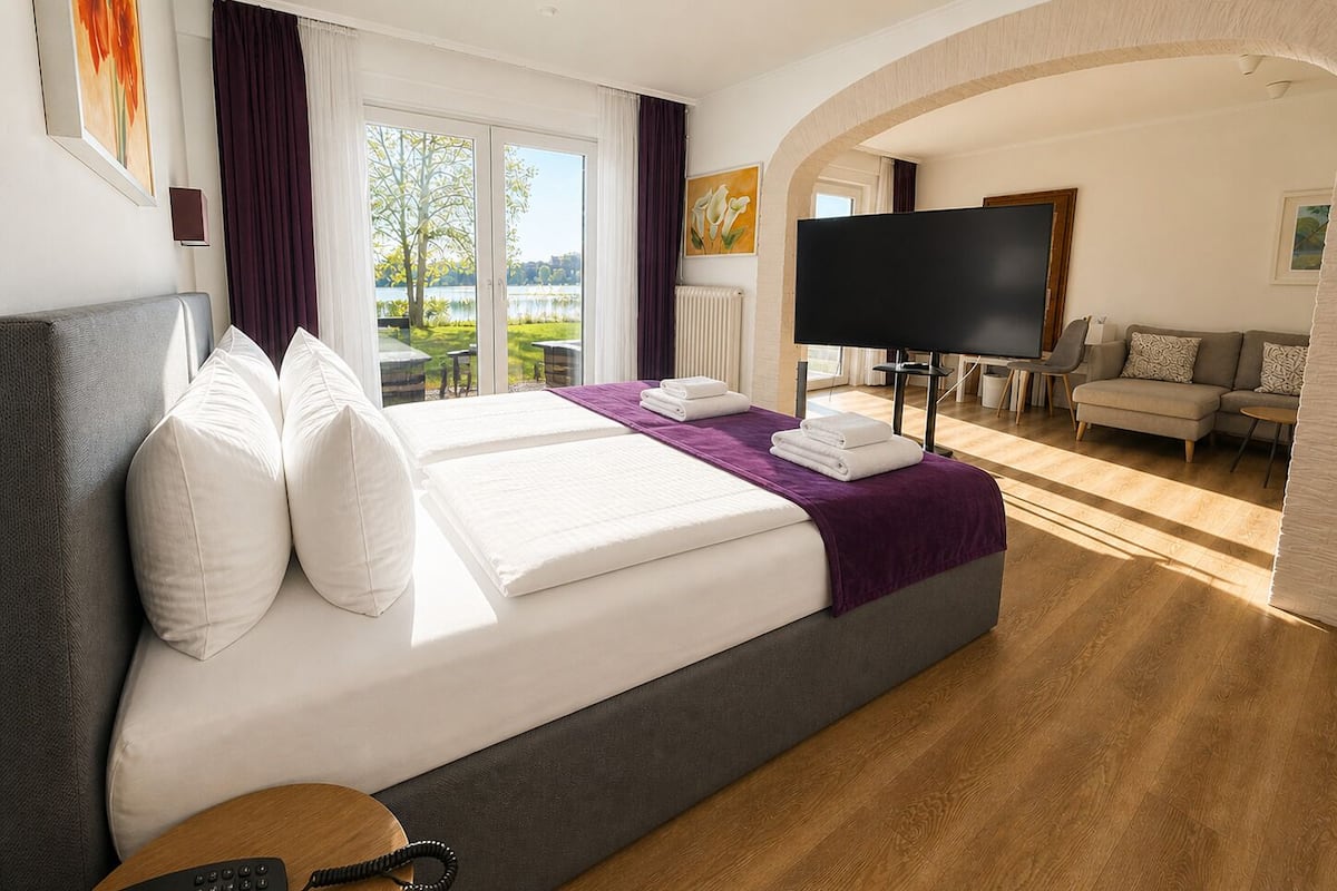 Quarto de hotel com uma cama grande, televisão, sofá e vista para um lago.