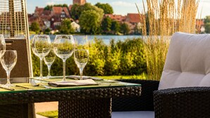 A dining table with four wine glasses, a green placemat, and cutlery, overlooking a body of water with buildings in the background. - Seehotel Schwanenhof (Moelln)