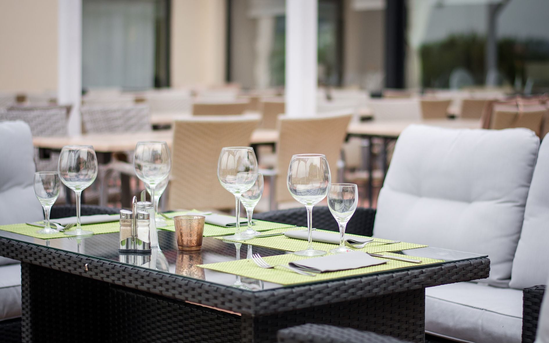 Une table à manger dressée avec des verres à vin, des couverts et une bougie, entourée de chaises en osier et d’un canapé blanc, le tout installé dans un espace extérieur.