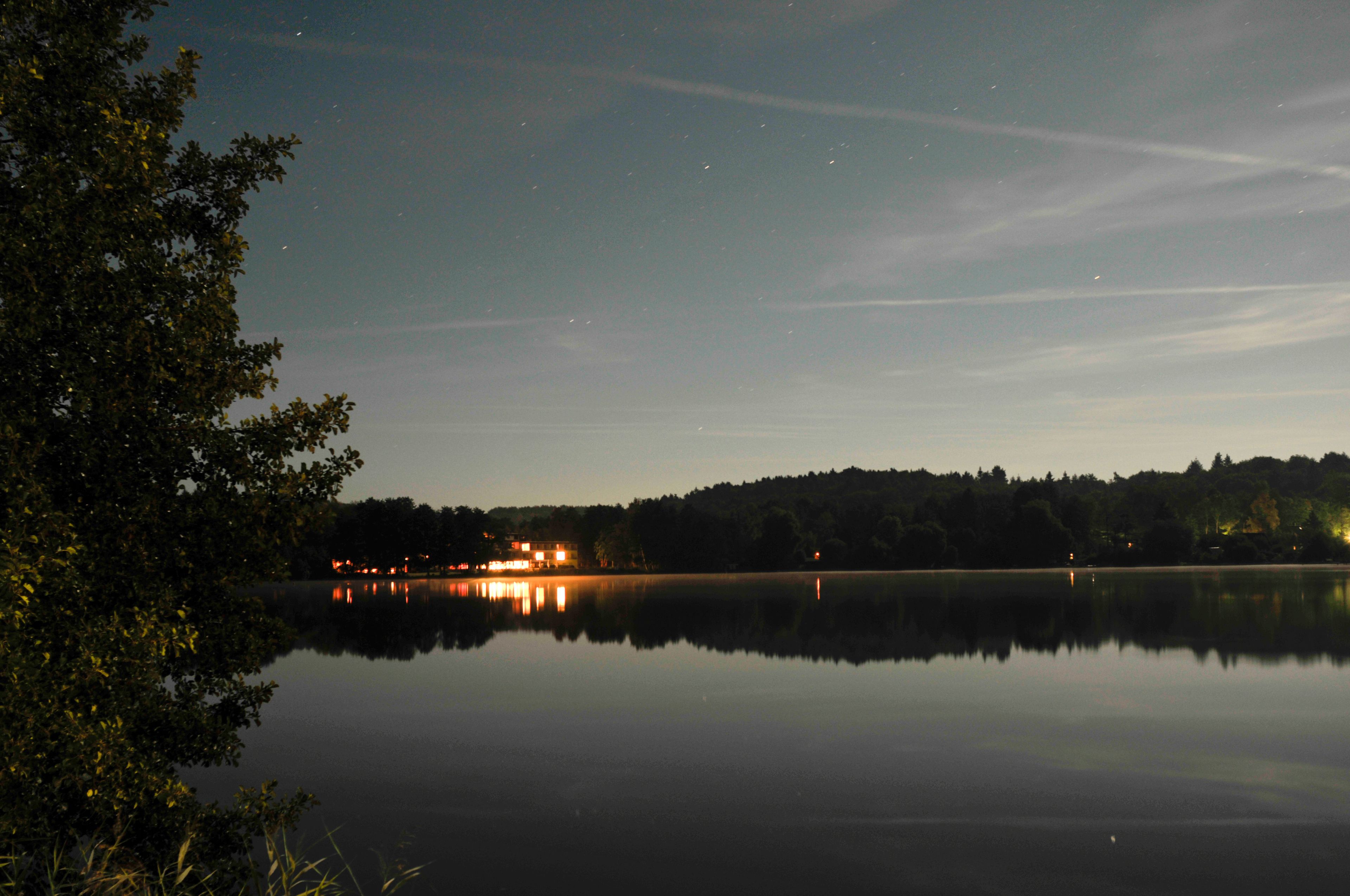 A calm lake at dusk with reflections of lights on the water, surrounded by trees and a distant hill with a building.