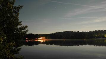 A calm lake at dusk with reflections of lights on the water, surrounded by trees and a distant hill with a building.