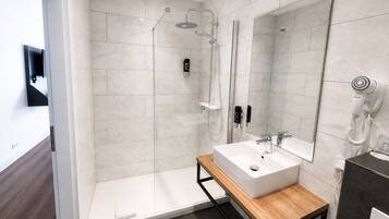 Un baño moderno con un espejo grande, un tocador de madera, una ducha con cabezal de ducha manual y un secador de pelo montado en la pared.