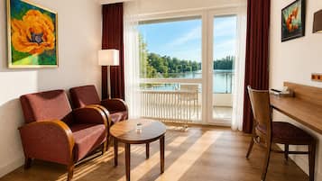 A hotel room with a sofa, chairs, a table, a lamp, a painting, a view of a lake, and a wooden desk.