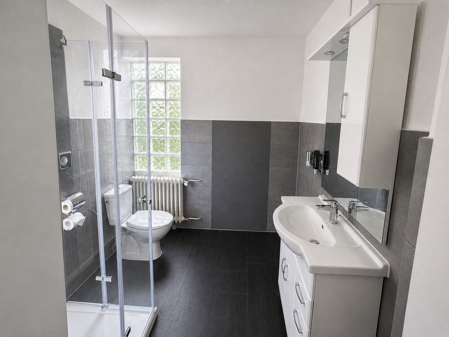 Een moderne badkamer met een douchecabine van glas, een toilet, een radiator en een wastafel met spiegel.