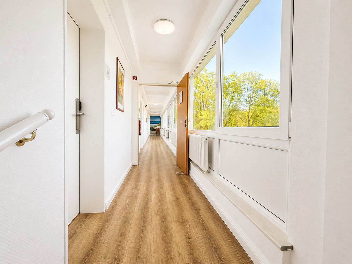 A hallway with wooden flooring, white walls, and a window with a view of trees.