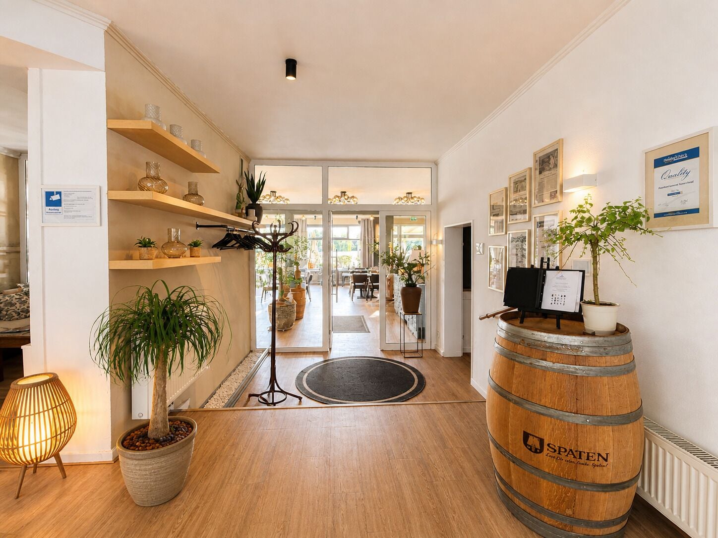 A hotel lobby with wooden flooring, a reception desk, a potted plant, a large barrel, and shelves with decorative items.