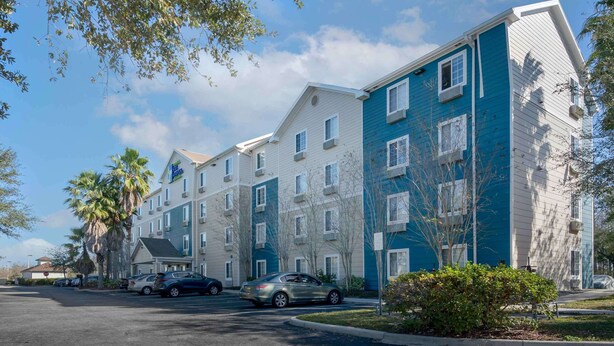Un hôtel à plusieurs étages, doté d’un design moderne, avec une façade aux couleurs variées, allant du blanc au bleu, et un parking où sont stationnées plusieurs voitures.