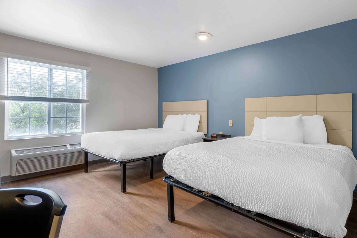 A hotel room with two single beds, a window with blinds, a heater, and a desk with a chair.