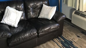 Un sofá de cuero negro con dos almohadas blancas frente a una pared azul, una lámpara de pie y una ventana con cortinas.