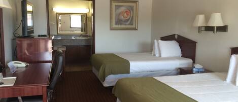 A hotel room with two beds, a desk with a chair, a telephone, a mirror, and a bathroom with a sink and mirror.