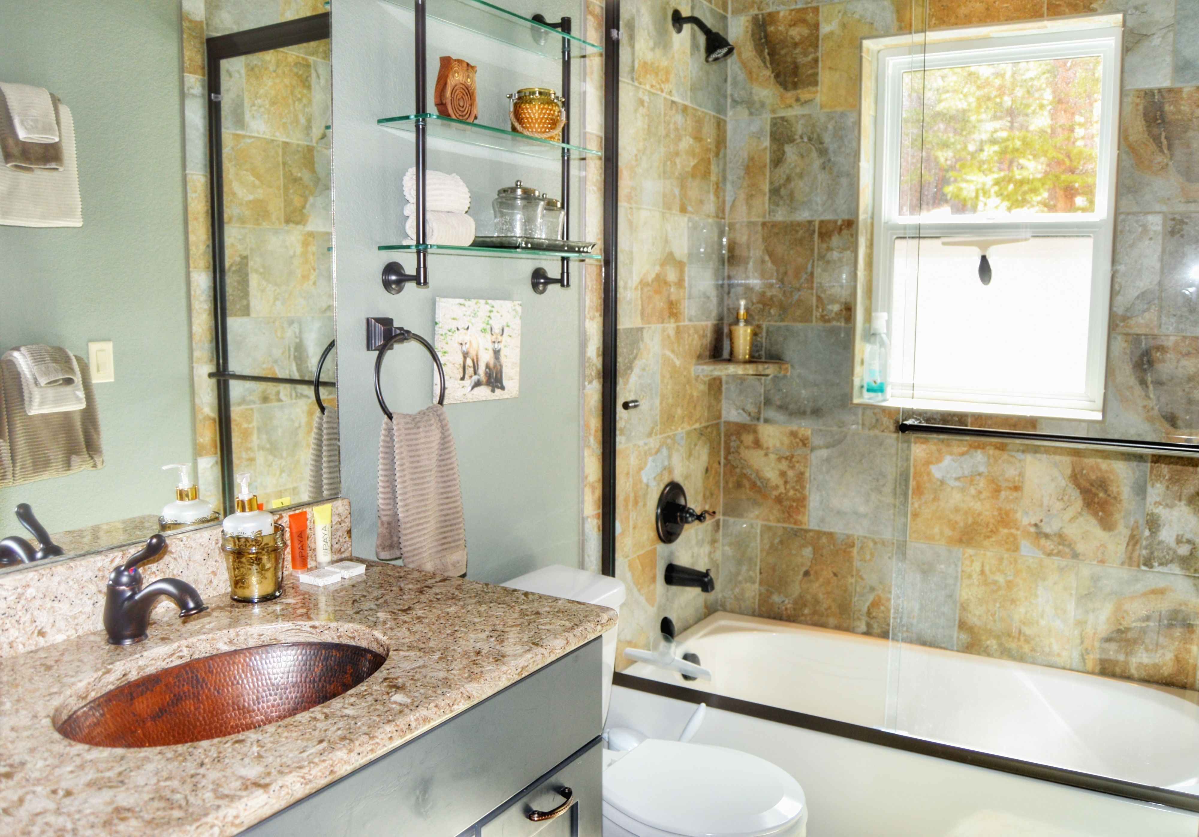 Une salle de bain avec un lavabo sur un plan de travail en granit, des toilettes blanches, une douche en verre et une fenêtre donnant sur les arbres.