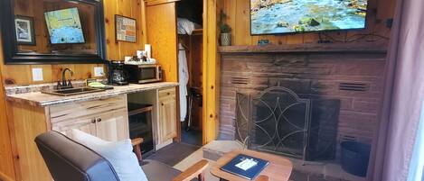 Ein gemütlicher Raum mit Kamin, einem an der Wand befestigten Fernseher, einer Küchenzeile mit Spüle und Herd sowie einer komfortablen Sitzecke mit Tisch.