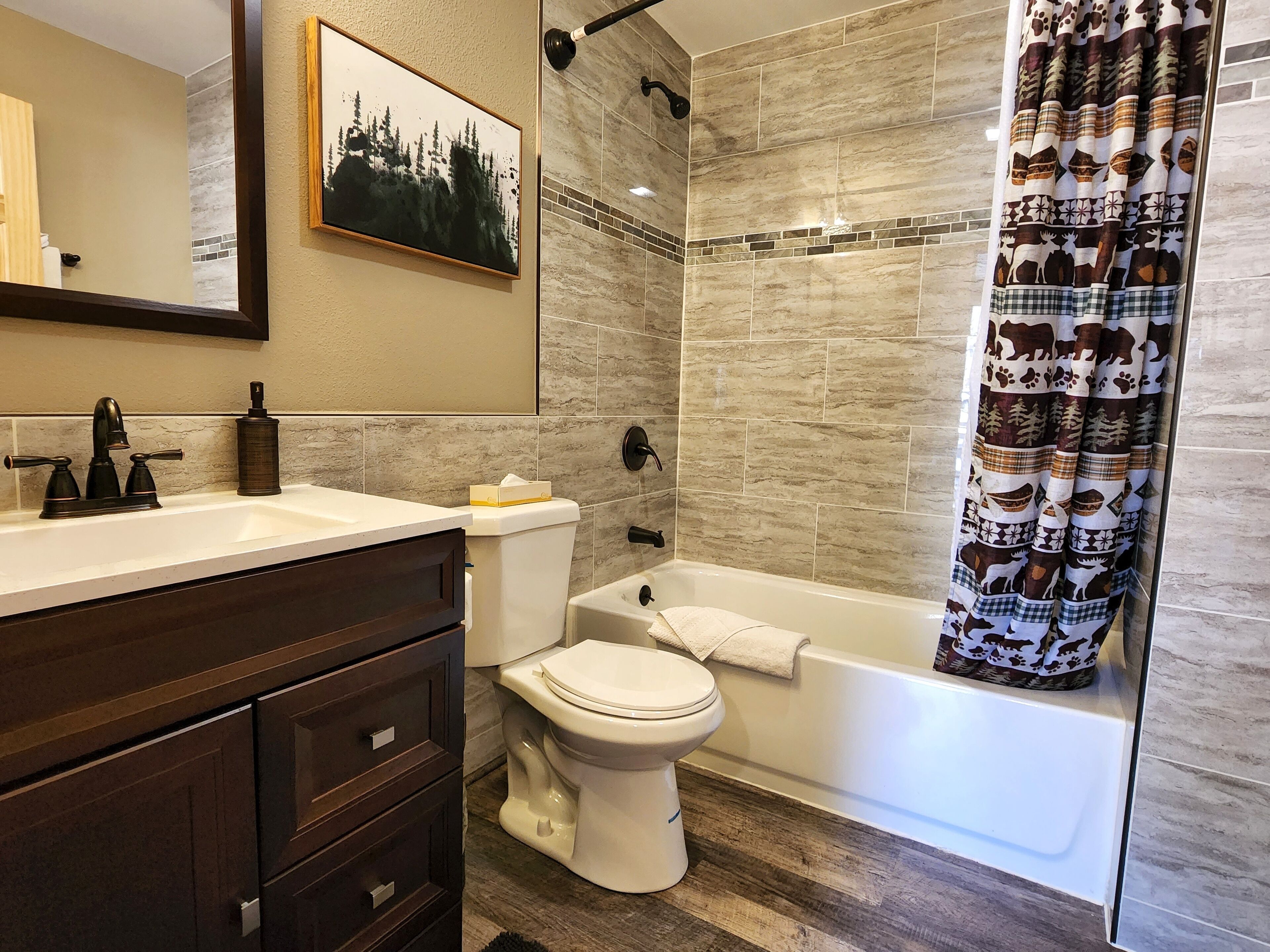 Ein Badezimmer mit Toilette, Badewanne, einer Duschvorhang mit Tiermotiven, einem Waschbecken mit dunklem Wasserhahn und einem gerahmten Bild an der Wand.
