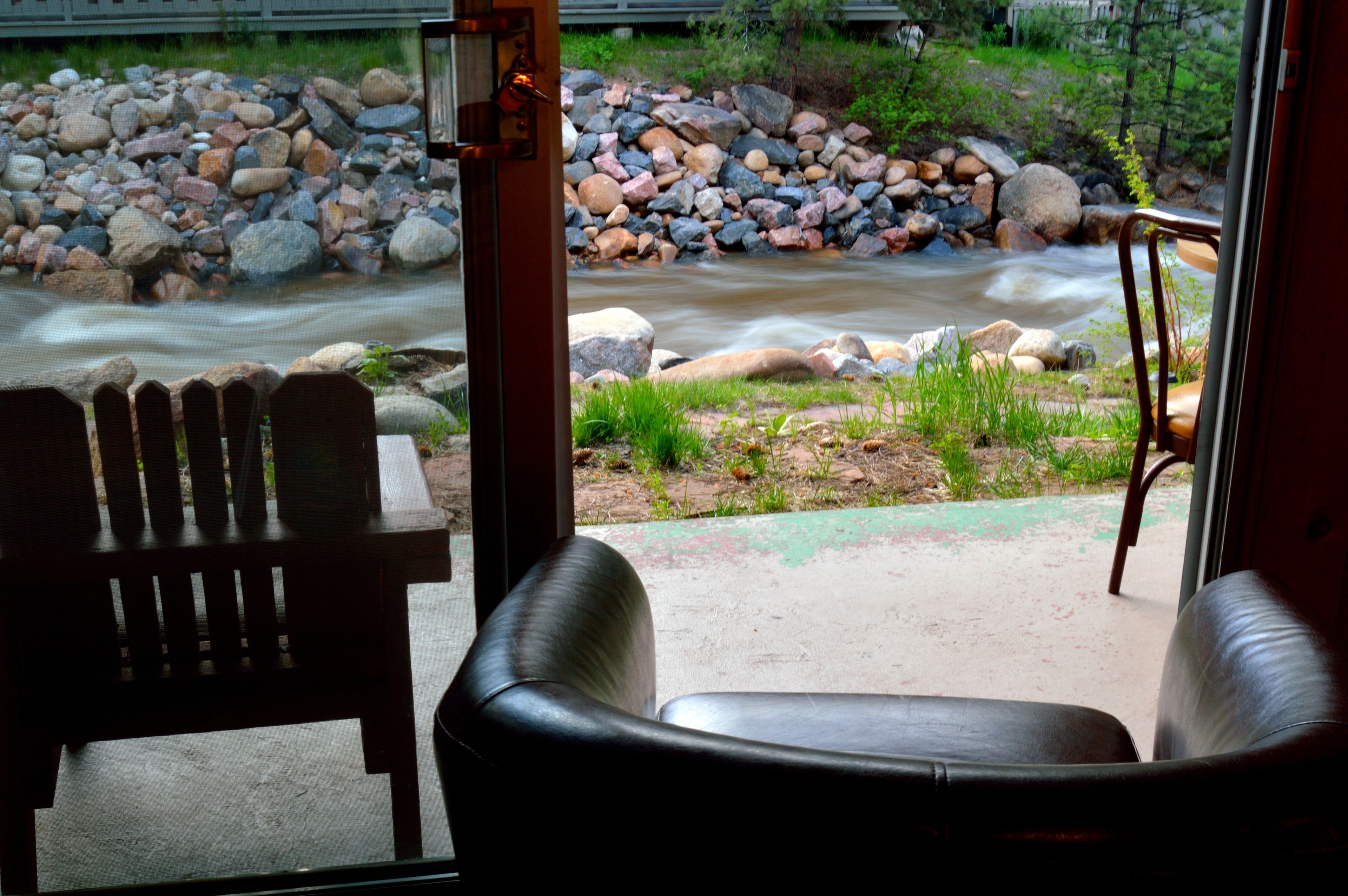A view of a river with rocks and greenery, a wooden bench, and a chair inside a building.