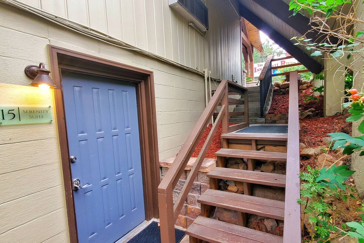 Une porte bleue sur laquelle est apposée une plaque indiquant « Suite Sérénité, n° 15 », et un escalier en bois menant à une zone abritée d’où l’on peut admirer la vue sur une rivière.