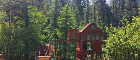 A wooden playhouse with a red roof and a swing set in a forested area.