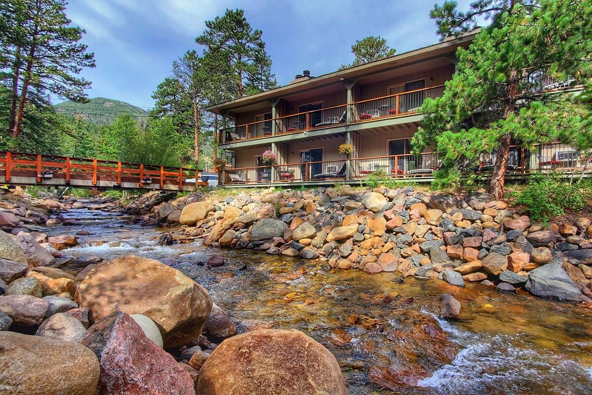 Un chalet avec un pont de pierre, un ruisseau et une chaîne de montagnes en arrière-plan.