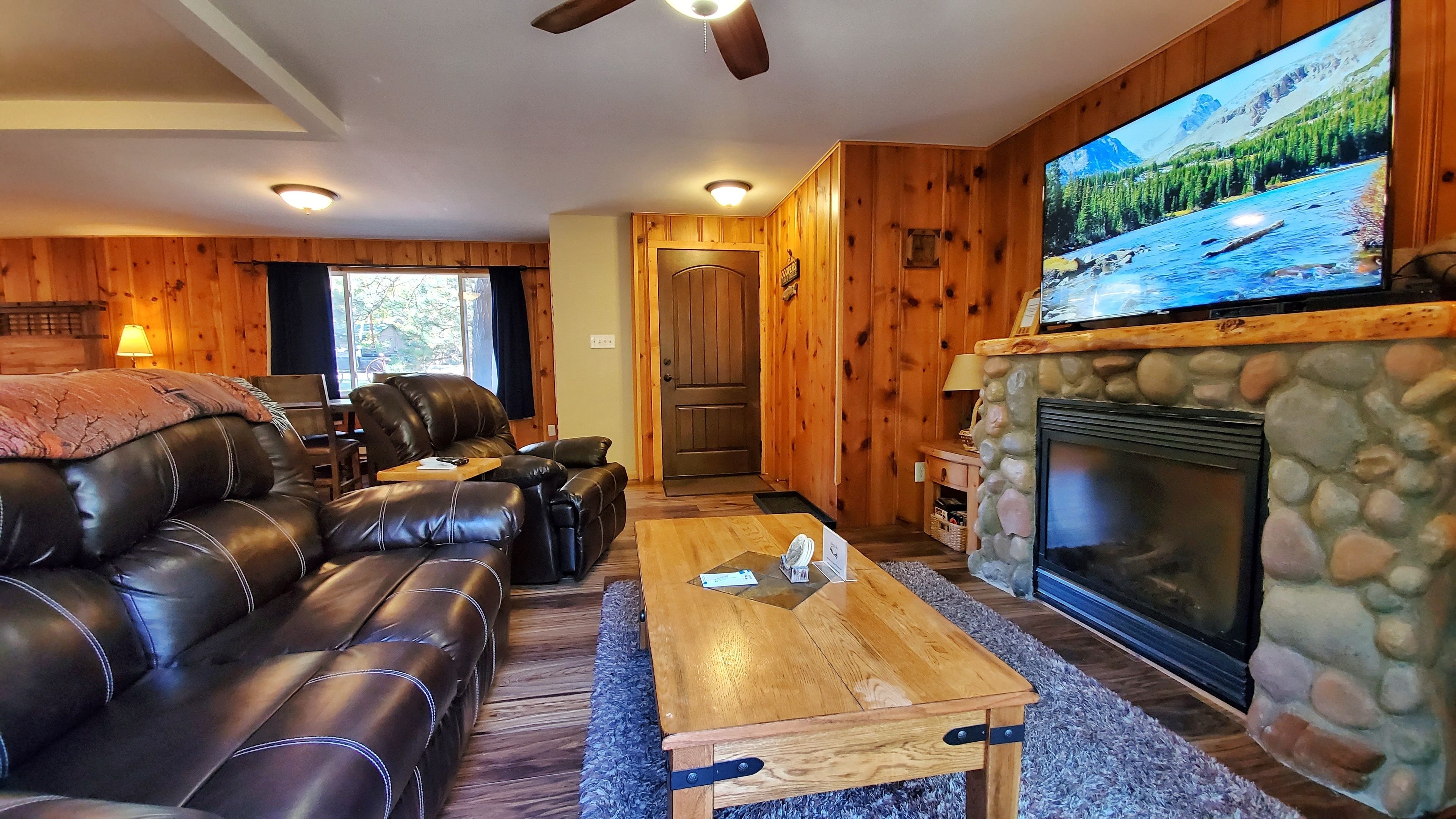 A cozy living room with a stone fireplace, leather couches, and a large TV screen displaying a scenic landscape.