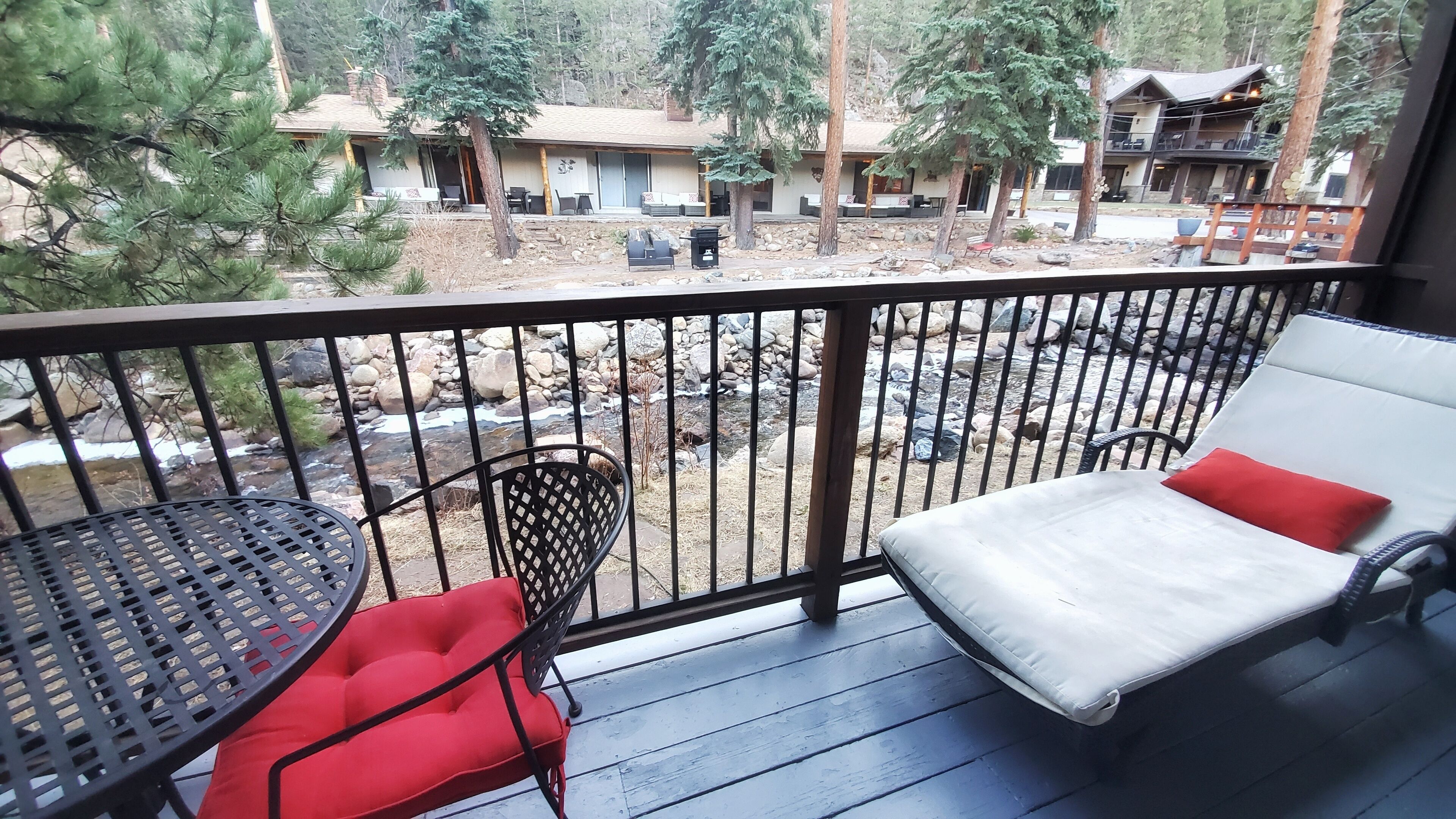 A balcony with a table and two chairs, overlooking a rocky area with a stream and trees.