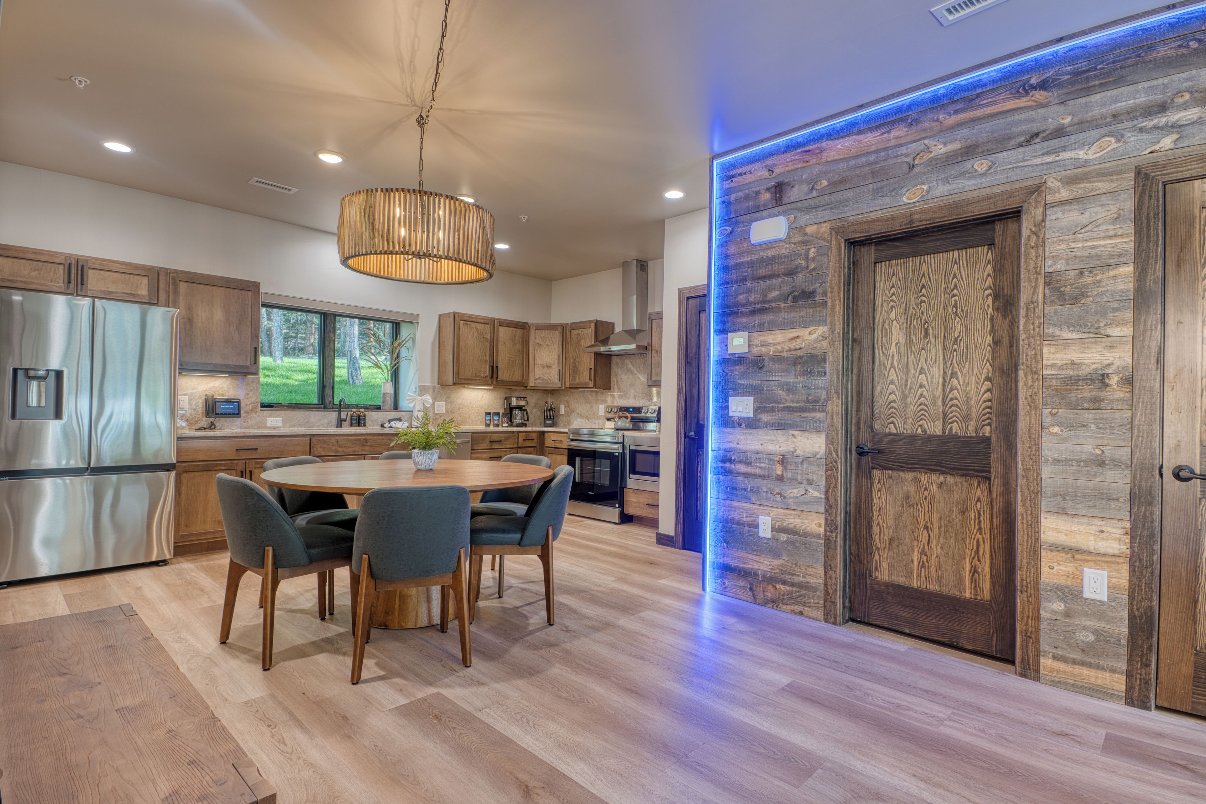 A modern kitchen with wooden cabinets, a stainless steel refrigerator, a dining table with chairs, and a large wooden door with a blue light.