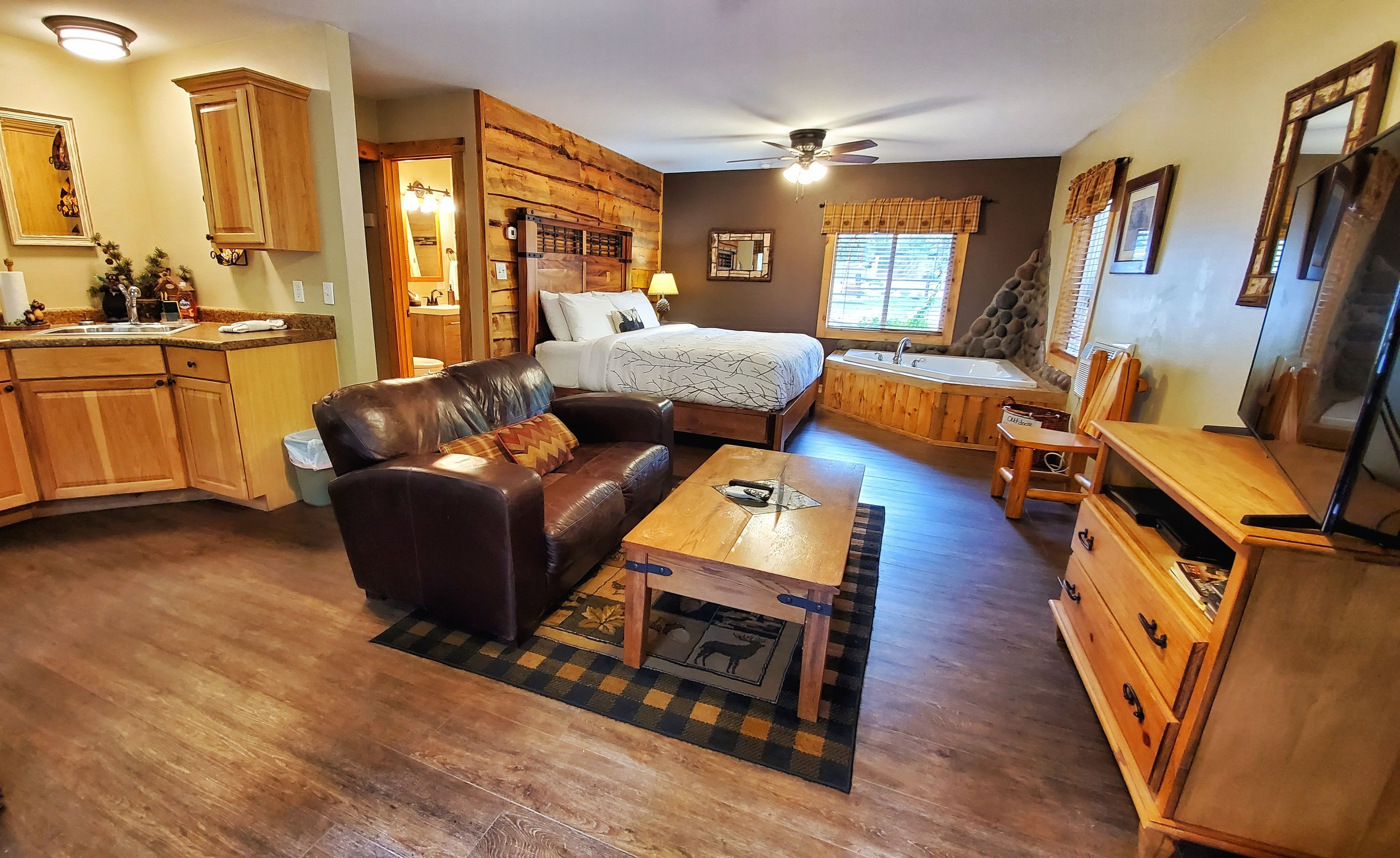 Una sala de estar acogedora con un sofá de cuero, una mesa de centro de madera y una cama con cabecera de madera. Hay una zona de cocina con armarios de madera y un lavabo.