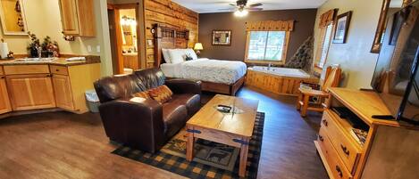 A cozy living room with a leather sofa, a wooden coffee table, and a bed with a wooden headboard. There is a kitchen area with wooden cabinets and a sink.
