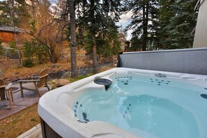 Un jacuzzi en un patio trasero con terraza de madera, sillas y un muro de piedra.