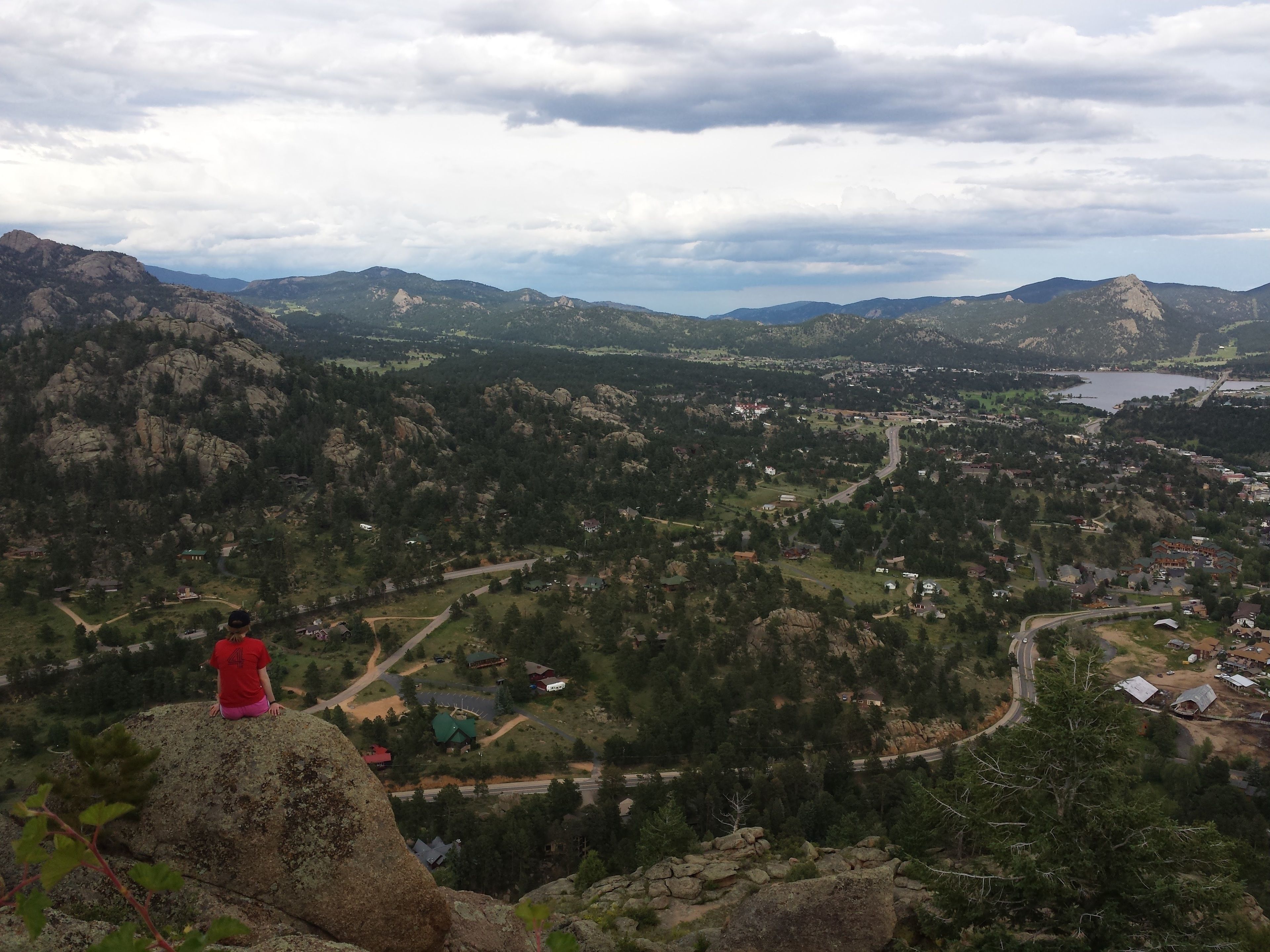 A person sitting on a rock overlooking a valley with houses and a lake.