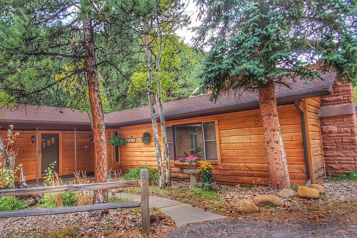 Un chalet rustique en rondins de bois, doté d’un porche, entouré d’arbres et bordé d’un chemin de gravier.