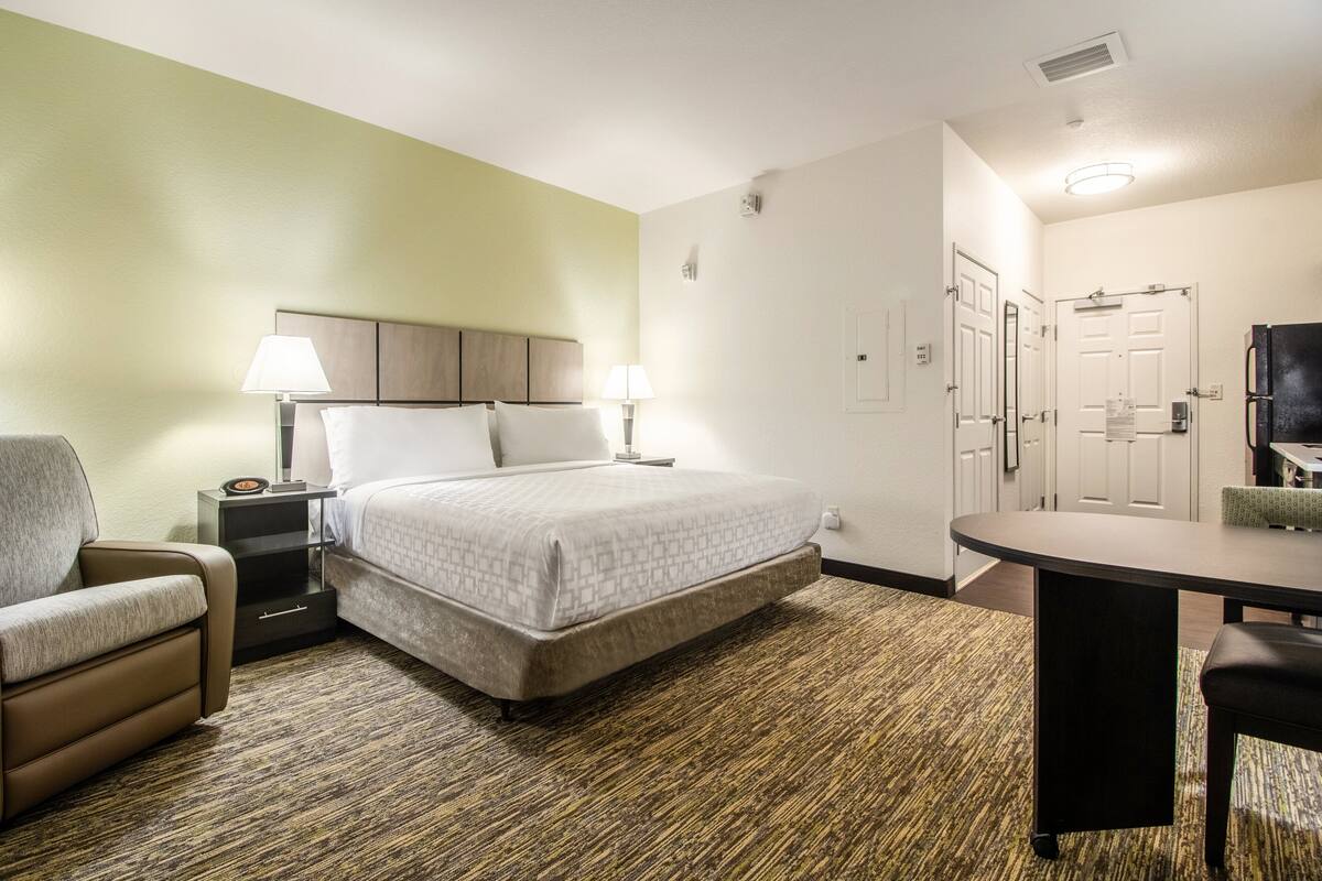 Una habitación de hotel con una cama, mesitas de noche, una silla, un escritorio y un refrigerador.