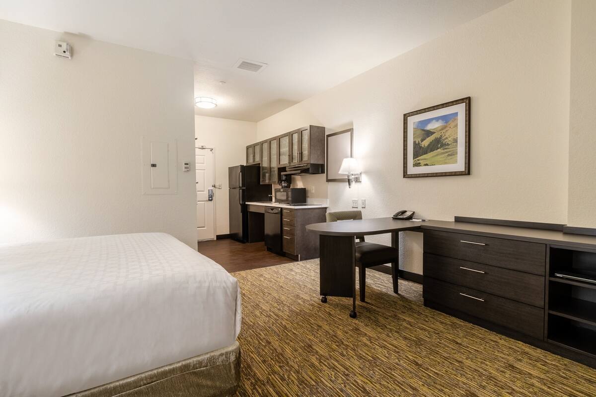 Habitación de hotel con cama, escritorio, silla, pequeña cocina y un cuadro en la pared.