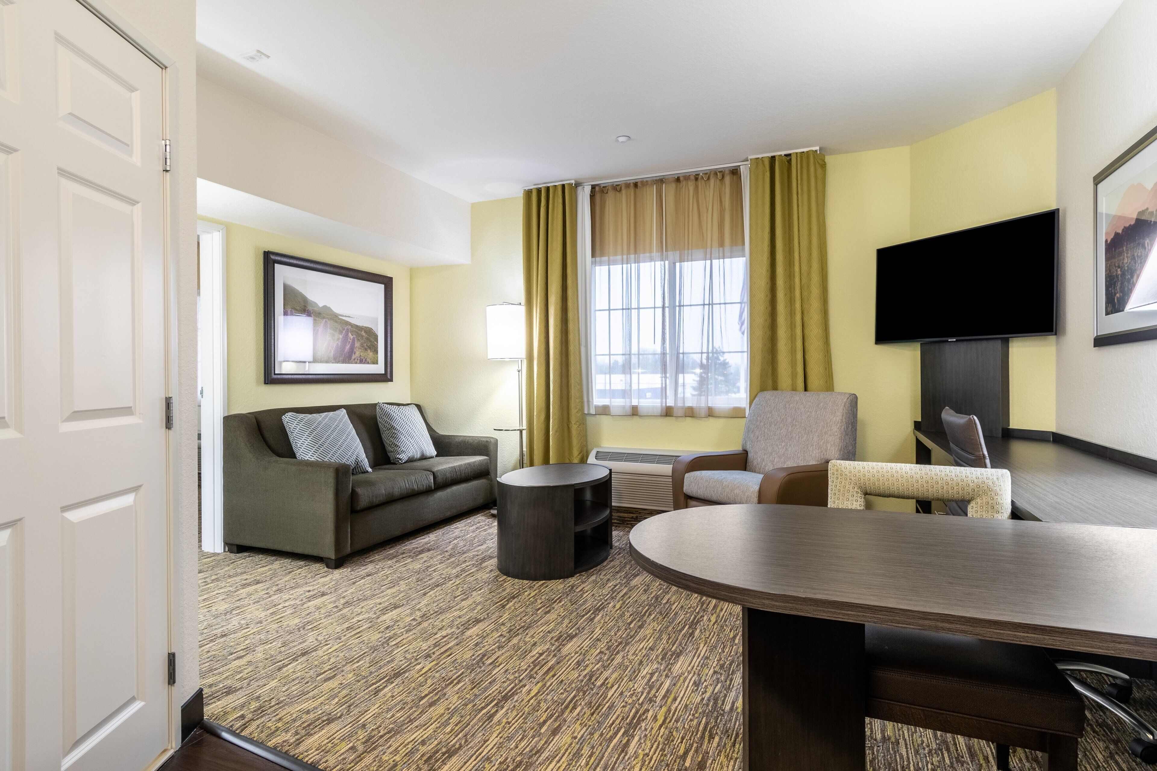 Habitación de hotel con sofá, sillón, mesa de centro, televisor de pantalla plana y un ventanal con cortinas.