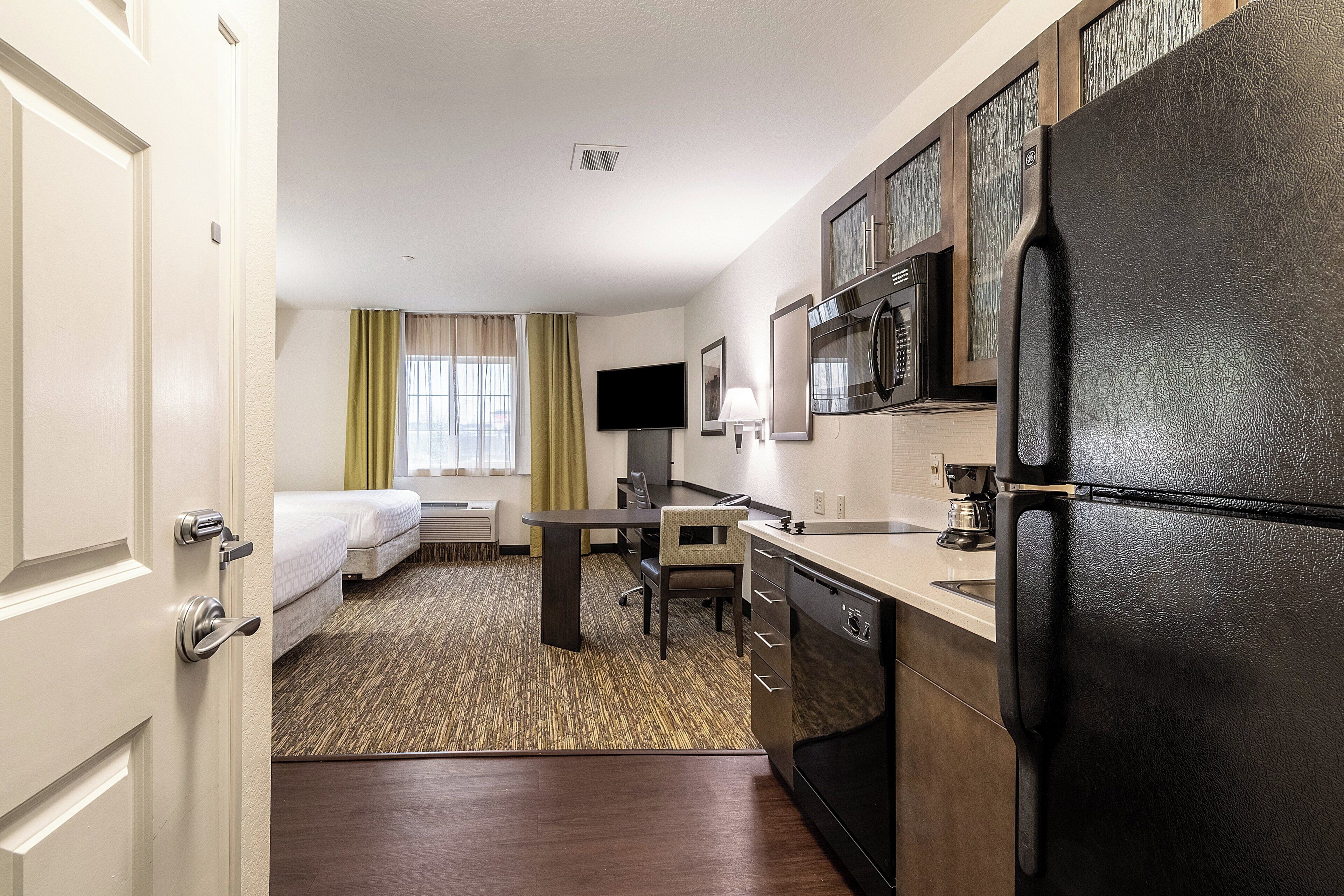 A hotel room with a bed, a desk, a chair, a refrigerator, a microwave, and a window with curtains.