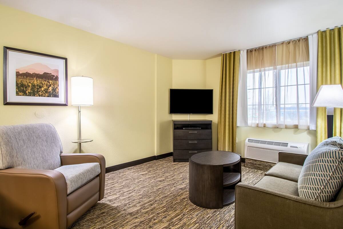 Habitación de hotel con sofá, una silla, televisión, lámpara y ventana con cortinas.