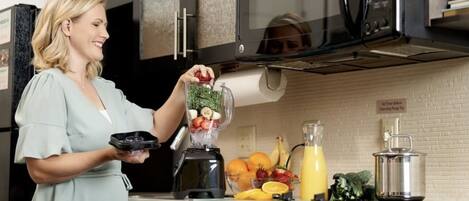 A kitchen with a microwave, a blender, and various fruits and vegetables on the counter.